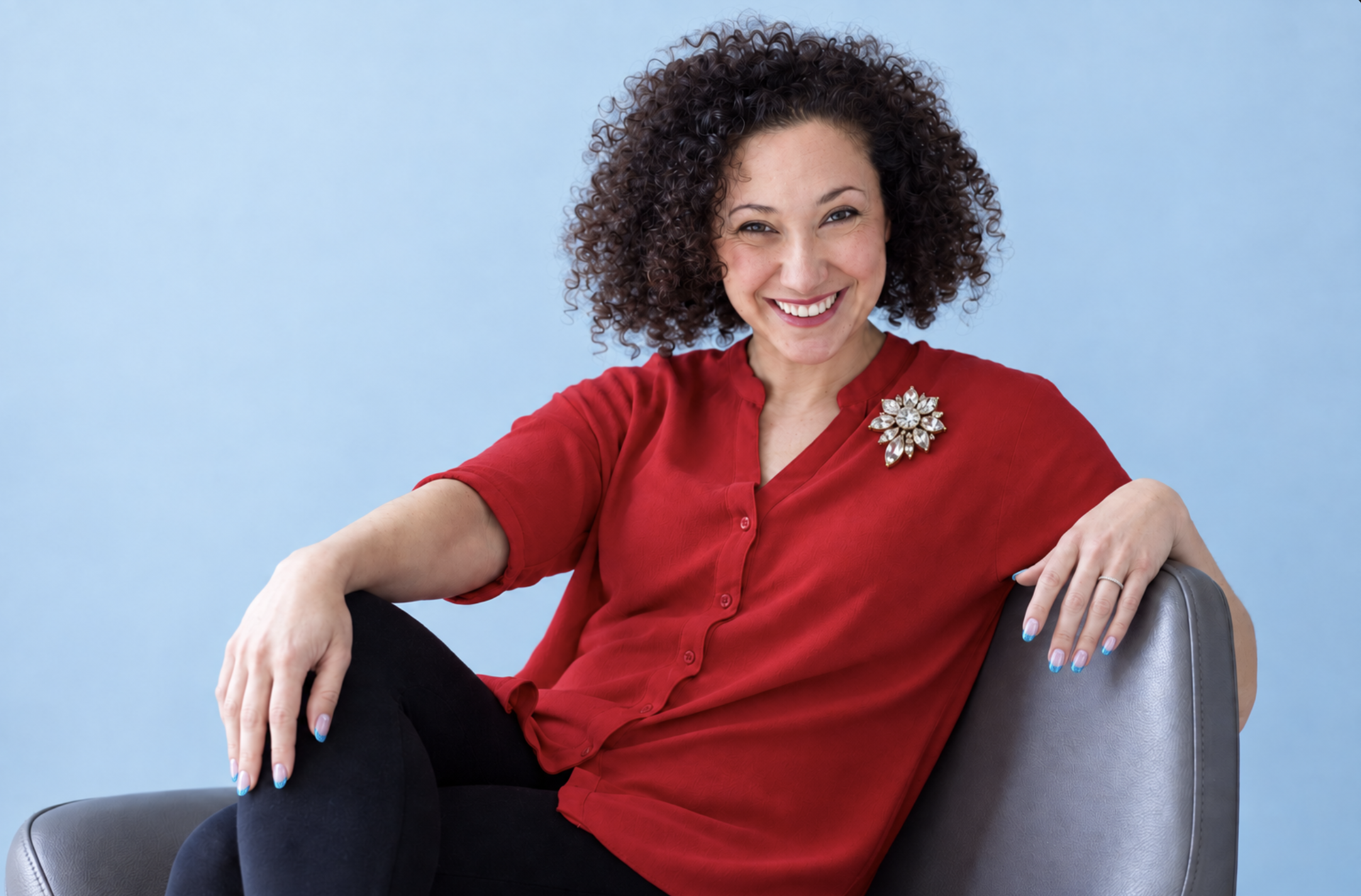 Nancy Abdalla seated in a cherry-red blouse with a crystal brooch against a powder-blue background
